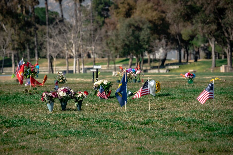 A wide view of a grassy memorial area at Altab Ali Park with numerous flower arrangements in various containers, including vases and wrapping, featuring a mix of fresh flowers such as pink and white c