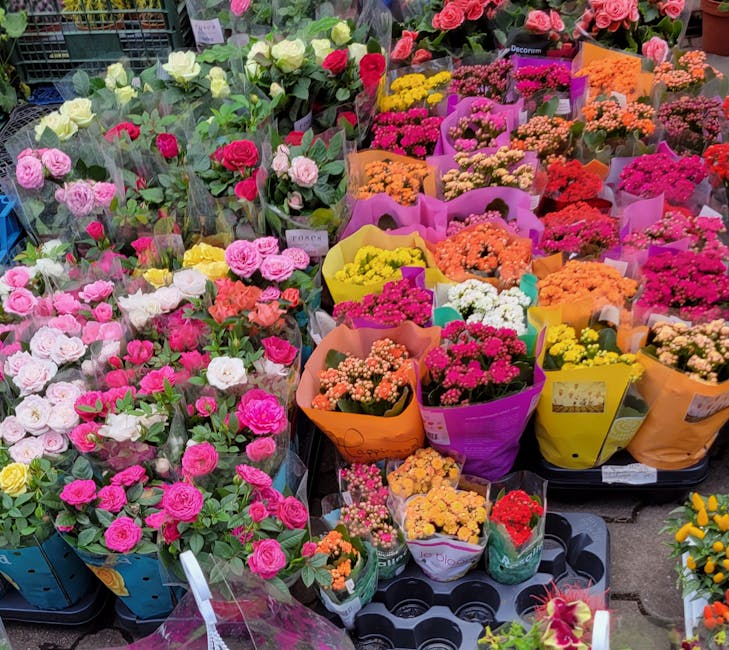 A vibrant display of fresh flowers at a flower market, featuring rows of pink, white, yellow, orange, and red blooms arranged in colorful wrapping paper. The flowers include roses, carnations, and sma
