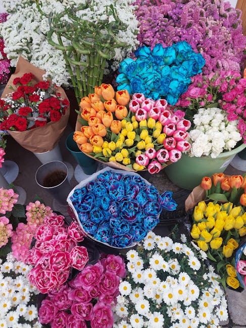 A vibrant assortment of fresh flowers arranged in various containers and bouquets. In the foreground, there are clusters of pink and red carnations, alongside white daisies with yellow centers. Behind