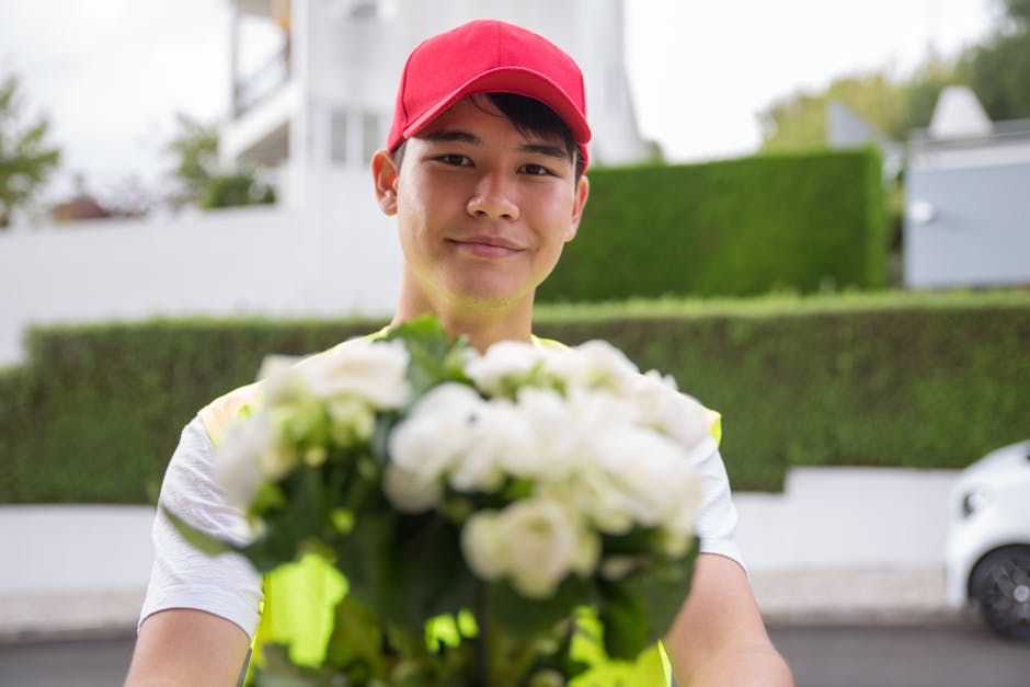 A delivery person wearing a red cap and a high-visibility vest is standing outdoors, holding a bouquet of fresh white flowers with green foliage, arranged in a rounded floral bouquet style. The flower