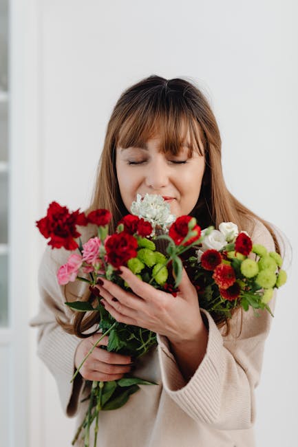 A woman with light brown hair and bangs, dressed in a cream-colored cardigan, is holding a vibrant bouquet of mixed flowers close to her face. The bouquet features red carnations, pink roses, white hy