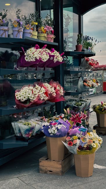 A display of various floral arrangements and bouquets arranged on metal shelves and wooden crates outside a flower shop. The flowers include white daisies, pink lilies, and purple orchids, wrapped in