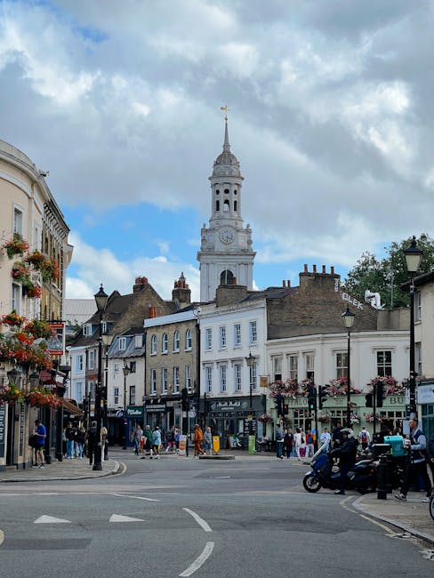 A view of a busy street scene in Whitechapel with historic terraced buildings, shops, and pedestrians. In the background, a tall, ornate clock tower with a spire rises against a partly cloudy sky, cas